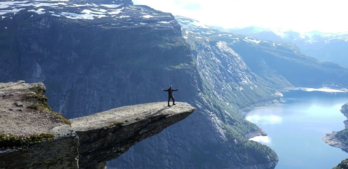 Manny standing with arms outstretched on a rock ledge high above a deep blue fjord